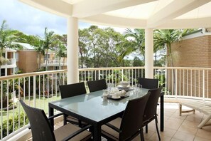 A balcony with a glass table set for dining, surrounded by wicker chairs, overlooking a garden with palm trees and a brick wall. - Macquarie Lodge Apartments (Sunshine Coast)