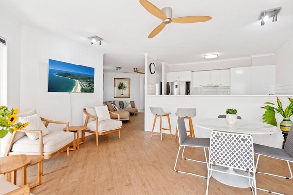 Ein modernes Wohnzimmer mit einer Sofagarnitur, einem Esstisch und Stühlen. Es gibt einen Deckenventilator, eine Küchenzeile mit weißen Schränken und ein gerahmtes Bild an der Wand.