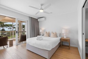 A modern hotel room with a large bed, a ceiling fan, a balcony with a view, and a small bedside table with a lamp. - Macquarie Lodge Apartments (Sunshine Coast)