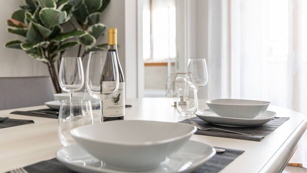 Uma mesa de jantar posta com pratos, tigelas e copos de vinho brancos, uma garrafa de vinho e um vaso de planta.