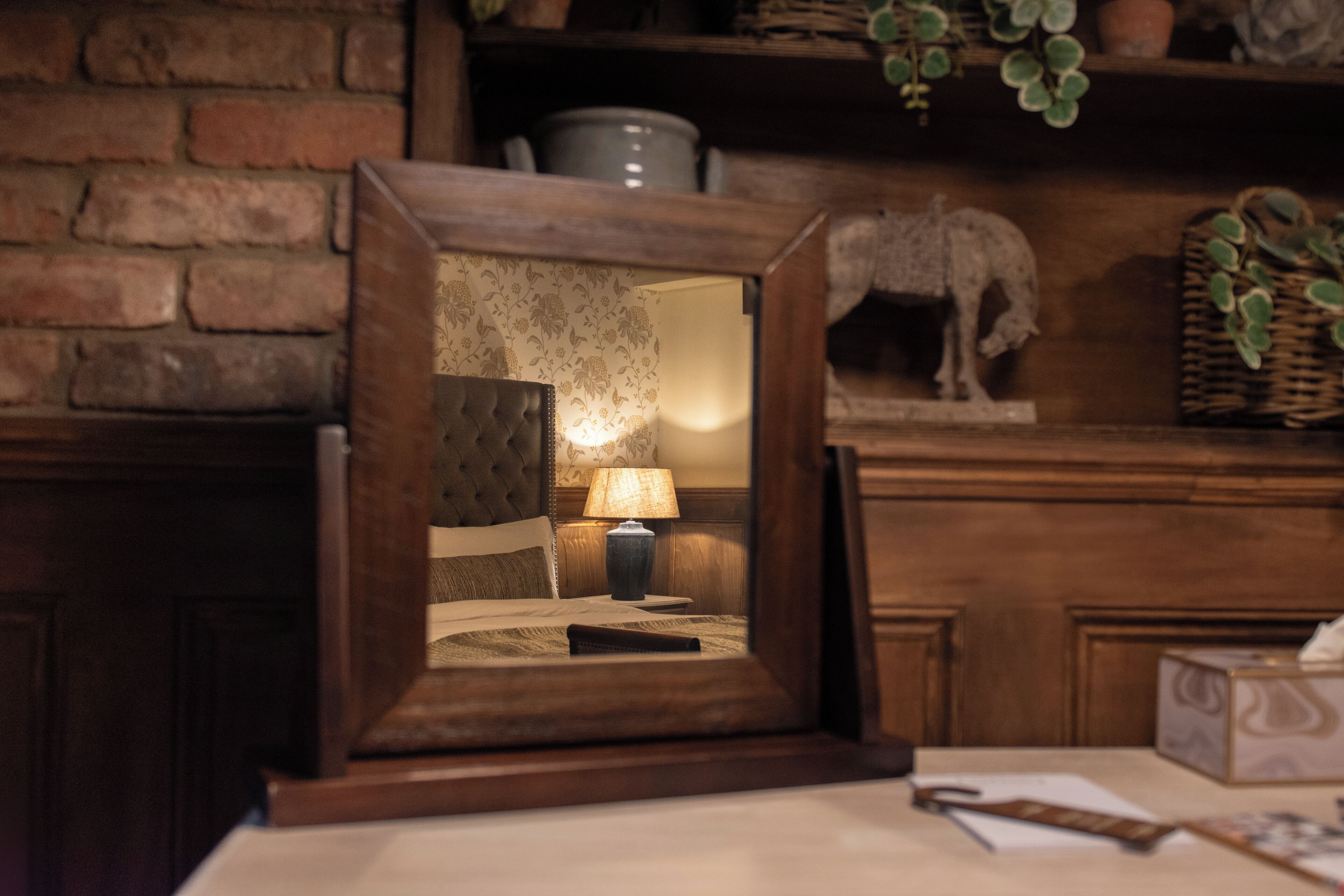 A framed mirror reflecting a bedroom with a bed, a lamp, and a nightstand.