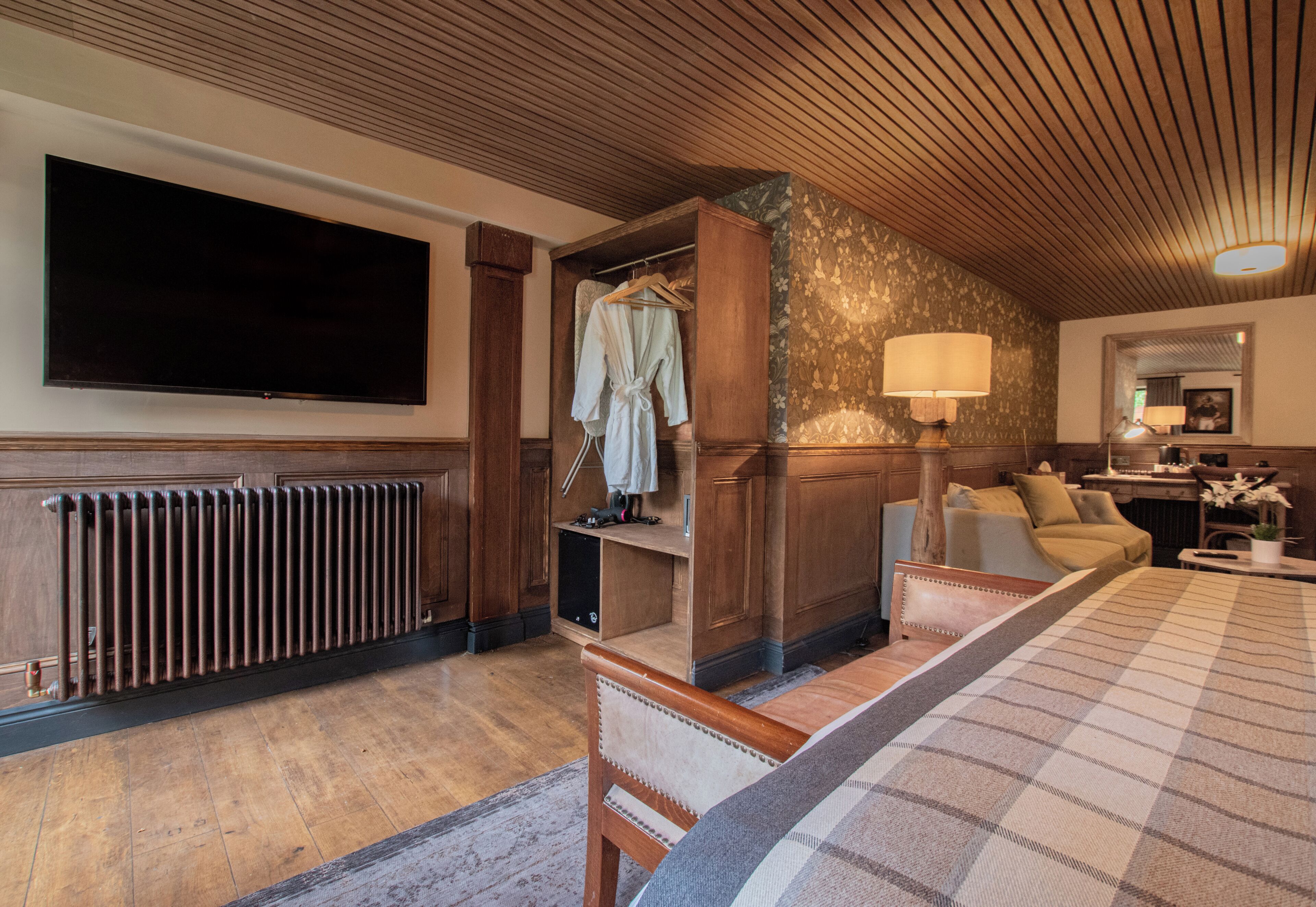 Ett hotellrum med en stor platt-TV, ett träpanelat tak, en säng med ett rutigt överkast, en sittgrupp med soffa och fåtöljer samt ett badrum med spegel och handduksställ.