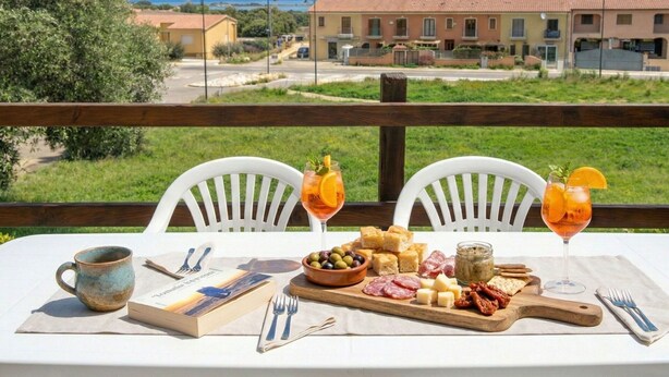Une table dressée avec un livre, une tasse, une planche en bois garnie de fromage, d’olives et de charcuterie, et deux verres d’une boisson aux agrumes, le tout placé devant un paysage pittoresque avec des bâtiments et de la verdure.