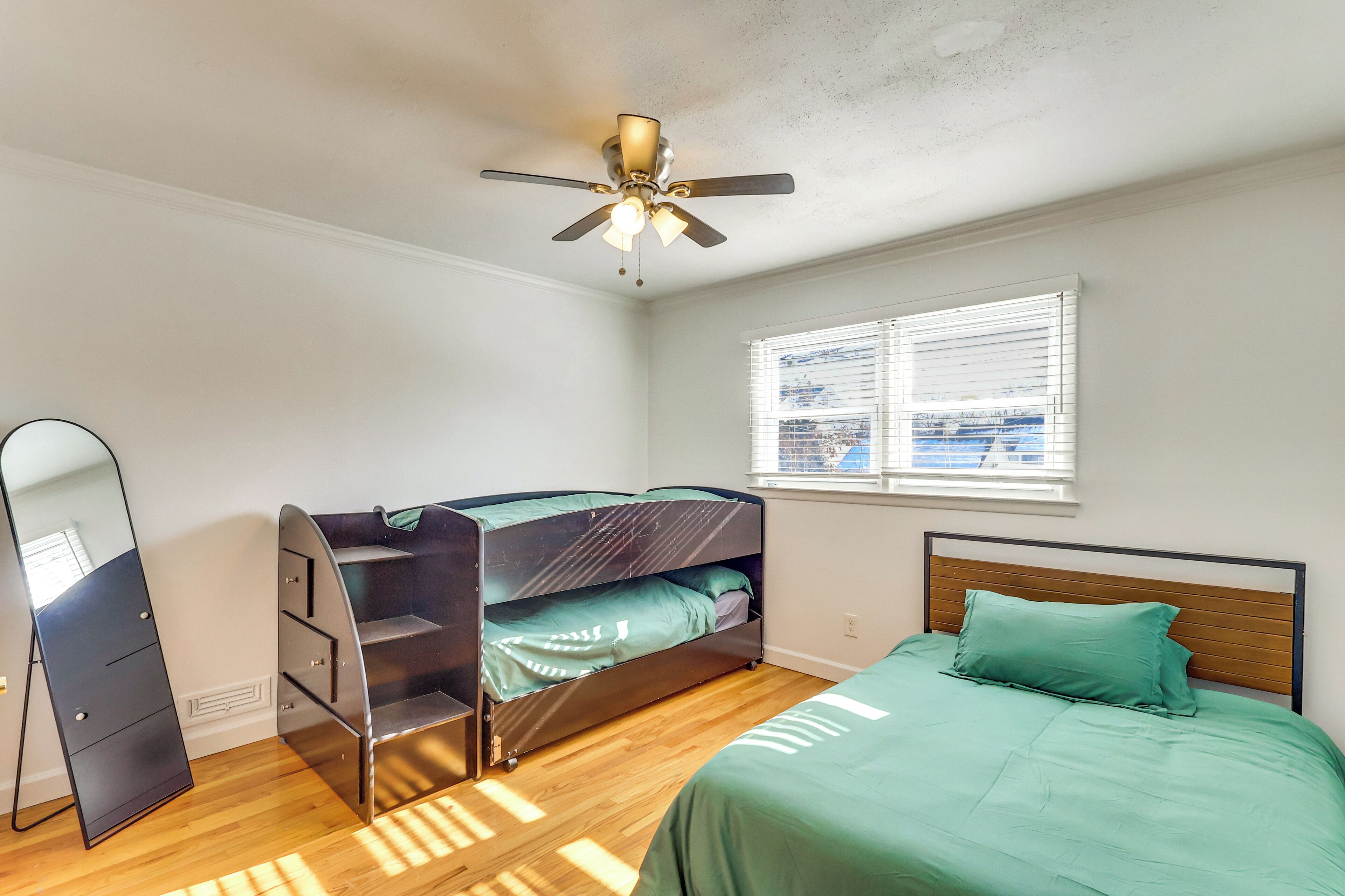Une chambre avec un lit superposé, un lit simple, un ventilateur de plafond, une fenêtre avec des stores et un miroir.