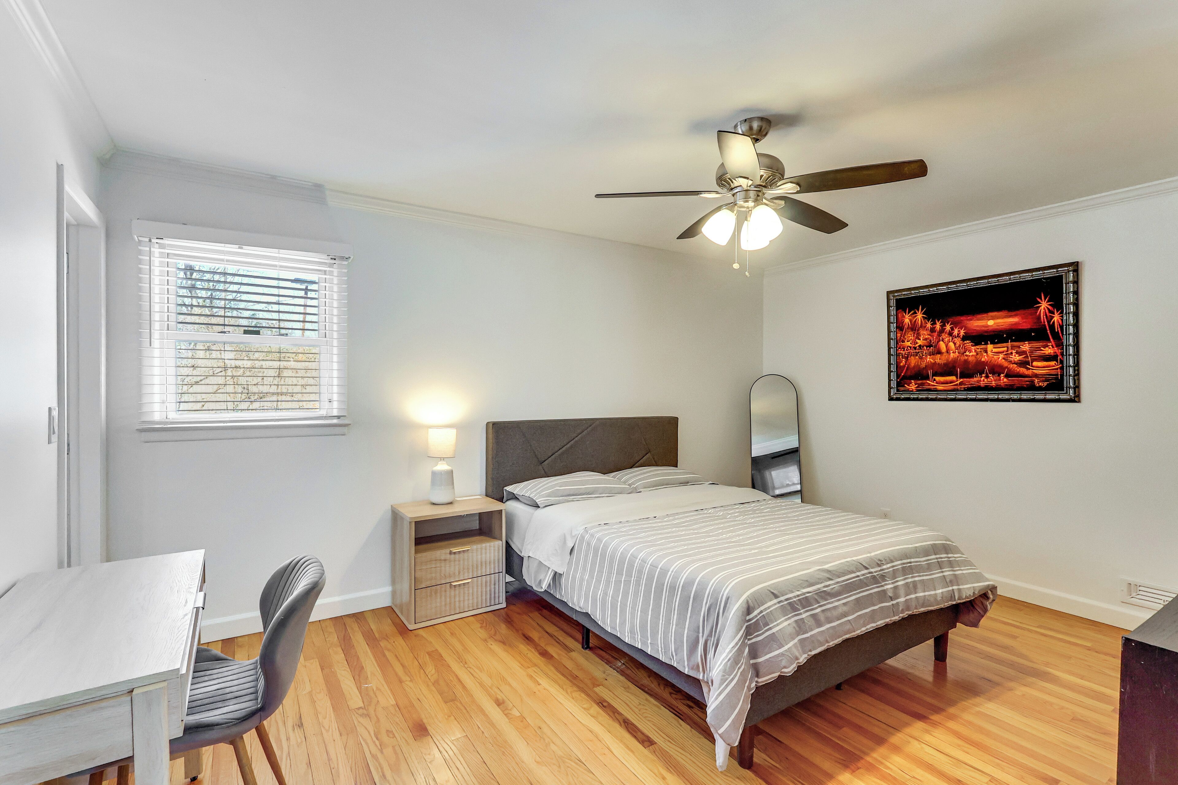 Une chambre à coucher comprenant un lit, une table de chevet, une chaise, une fenêtre, un ventilateur de plafond et un tableau encadré accroché au mur.