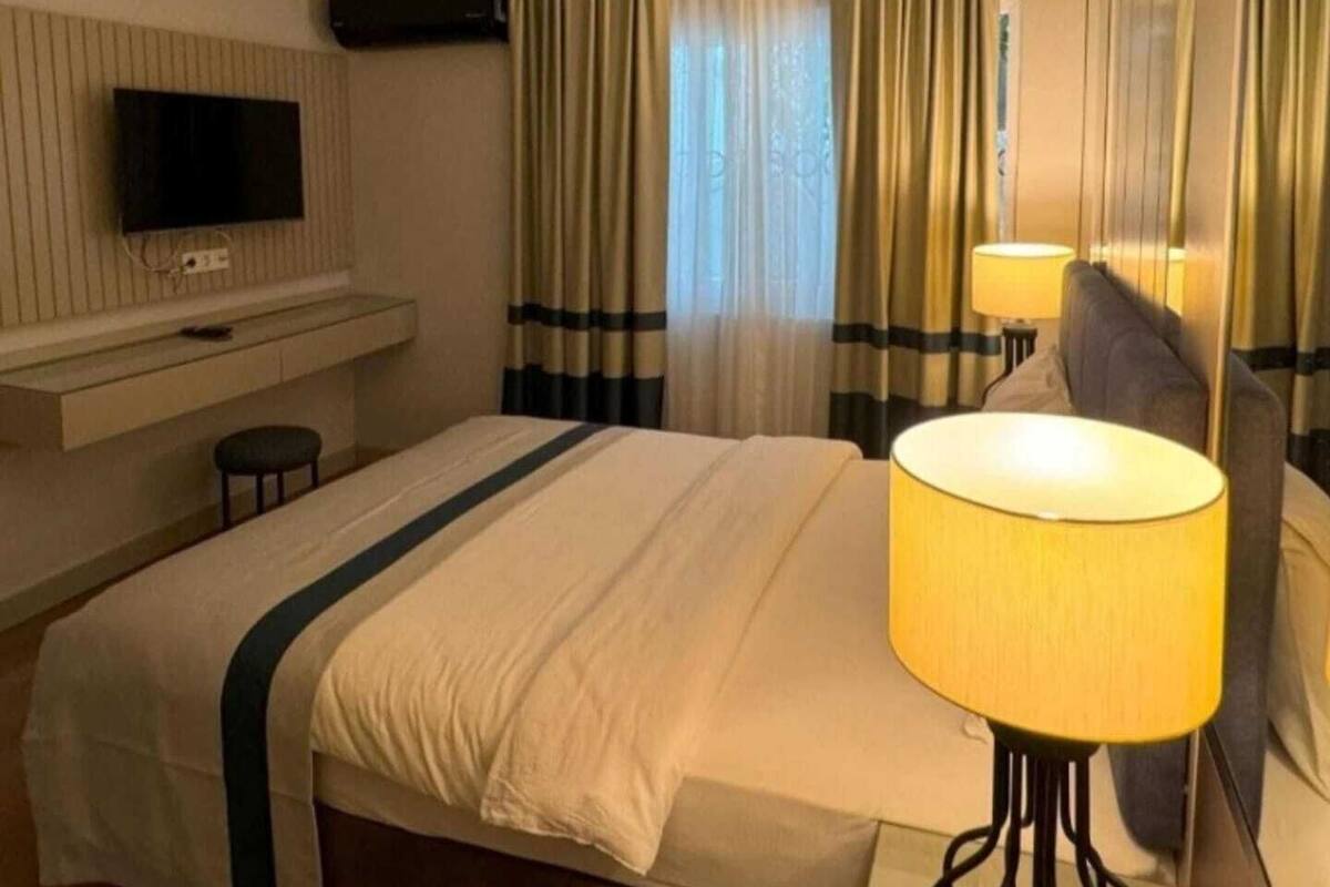 Ein Hotelzimmer mit einem Bett, einem an der Wand montierten Fernseher, einer Nachttischlampe und einem Fenster mit Vorhängen.