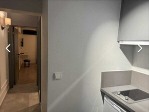 A modern hotel room with a kitchenette, a door leading to a hallway, and a small stool. - EBRU EVLERI ICMELER MERKEZE PLAJA YAKIN (Marmaris)