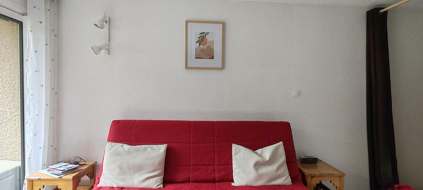 Een rode bank met twee kussens, een klein houten tafeltje met een boek, en een lijst met een foto aan de muur.