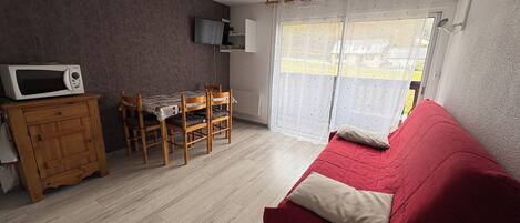 Una sala de estar con un sofá rojo, una mesa de comedor de madera y sillas, un microondas en un mueble, y un televisor montado en la pared.