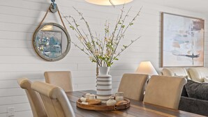 Ein Essbereich mit einem Holztisch, beigen Stühlen, einer Vase mit Zweigen, einem Spiegel und einem Gemälde an der Wand.