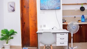 A room with a wooden door, a desk with a chair, a television, a fan, and a potted plant. - Marve 408 Stylish Studio Apartment (Mtwapa)