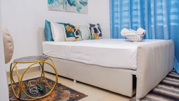 A bedroom with a bed, a small round table, a chair, and a window with blue curtains. - Marve 408 Stylish Studio Apartment (Mtwapa)