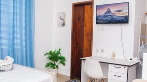 A hotel room with a bed, a desk with a chair, a potted plant, a wooden door, and a wall-mounted TV. - Marve 408 Stylish Studio Apartment (Mtwapa)