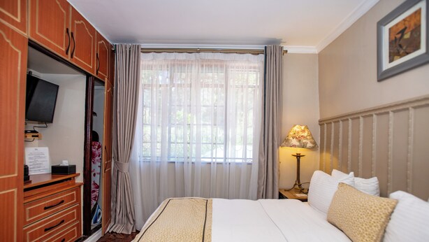 A hotel room with a bed, a television, a desk, and a window with curtains. - Standard Room in Gigiri near UN Nairobi, Cozy Stead Garden House (Nairobi)