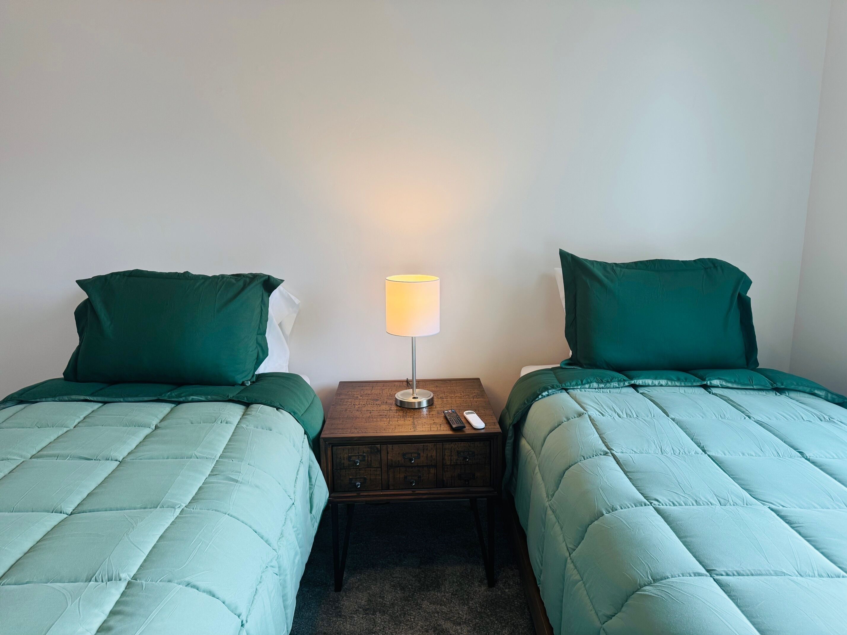 A twin room with two beds, each with a green duvet and pillow, a bedside table with a lamp, and a remote control on the table.