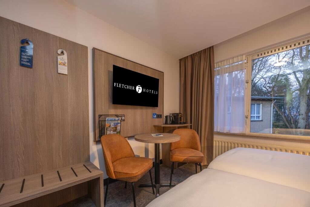 A hotel room with a bed, two armchairs, a small table, a TV mounted on the wall, and a window with curtains.