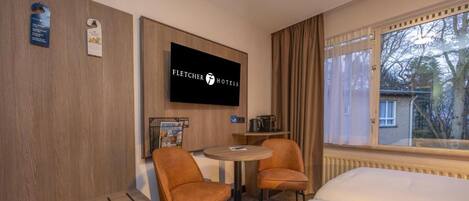 A hotel room with a bed, two armchairs, a small table, a TV mounted on the wall, and a window with curtains.
