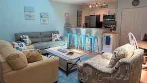 Una sala de estar con sofá, sillón, mesa de centro y una zona de cocina con refrigerador, microondas y taburetes de bar.