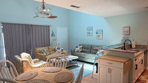 Una sala de estar con sofá, sillas, una mesa de comedor y una zona de cocina con pileta y alacenas.