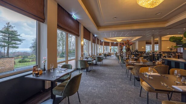 Un restaurant spacieux avec de hauts plafonds, de grandes fenêtres et un lustre. Les tables sont dressées avec des verres à vin et des couverts.