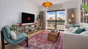 A modern living room with a flat-screen TV, a sofa, a coffee table, and a large window with a view of pyramids.