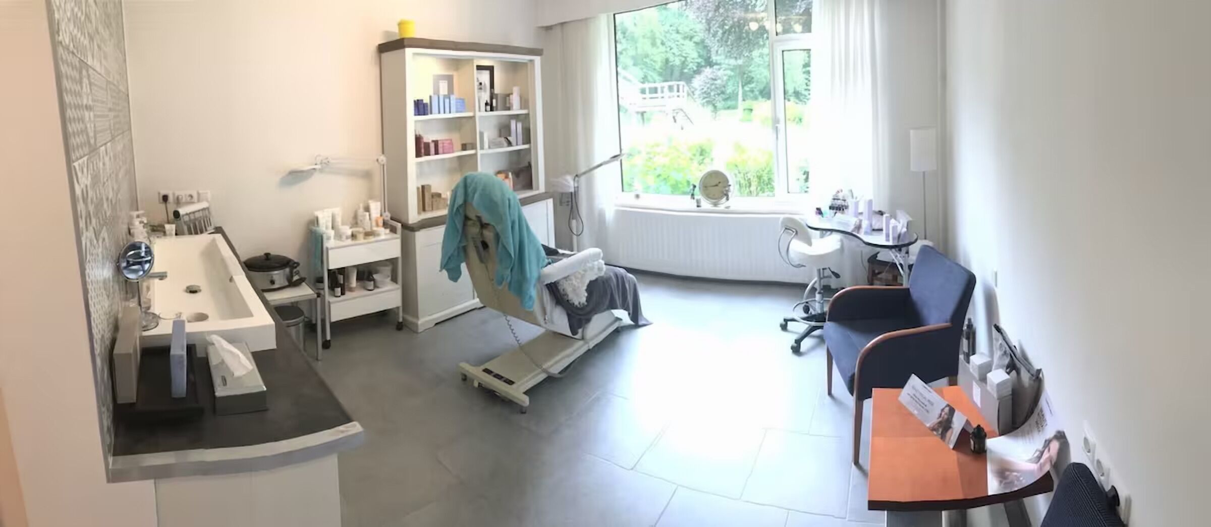 Eine moderne Zahnarztpraxis mit Empfangstrespo, Wartebereich mit Sitzgelegenheit, großem Fenster mit Blick auf Grünflächen und Regal mit diversen Gegenständen.