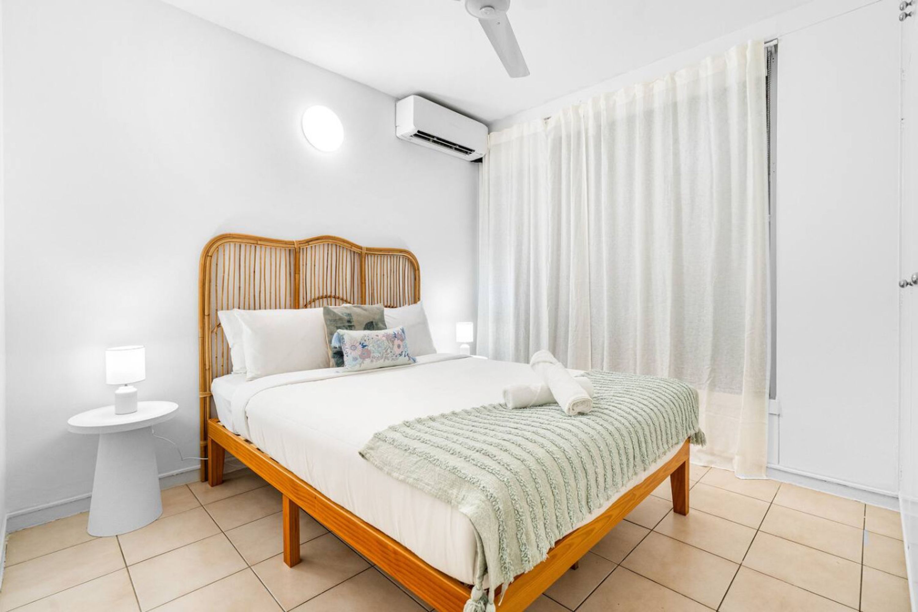 A neatly arranged bedroom with a wooden bed, a bedside table, an air conditioning unit, and a window with curtains.