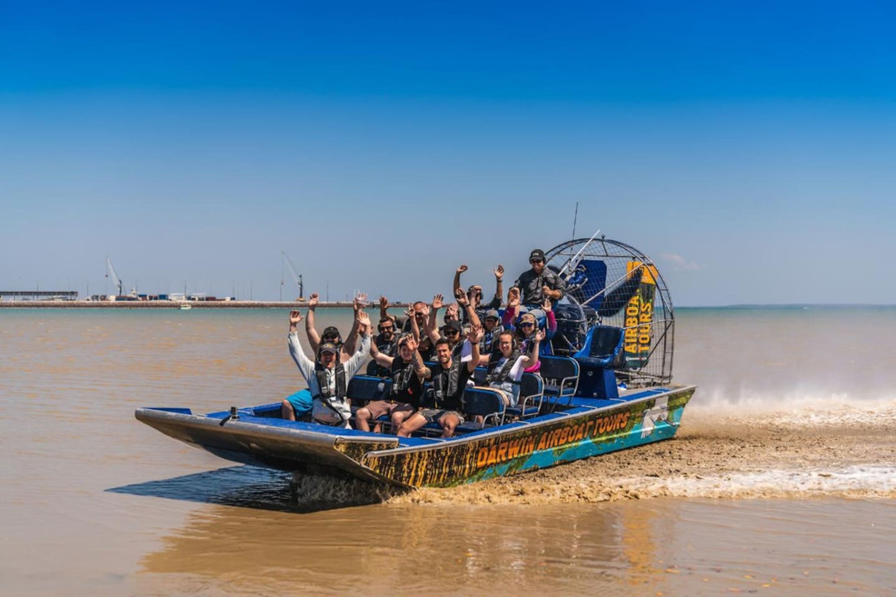A boat with passengers raising their hands, moving through a body of water with a clear sky in the background.