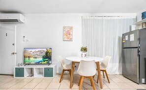 Cozinha moderna com geladeira embutida, mesa de jantar com cadeiras, TV de tela plana presa a um armário branco e uma janela com cortina branca.