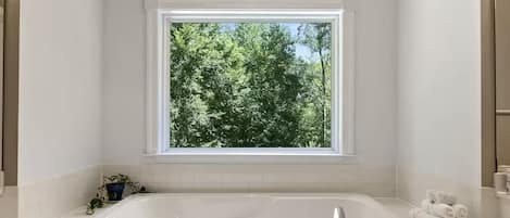 Un baño moderno con una bañera grande, una ventana con vistas a árboles y una planta en maceta sobre el mostrador.