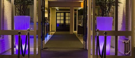 L’entrée d’un hôtel, avec une porte en verre, un chemin recouvert de moquette et des plantes en pot de chaque côté.