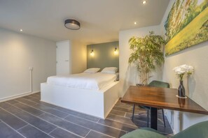 Ein modernes Schlafzimmer mit einem Bett, einem Esstisch, Stühlen, einer Topfpflanze und einem Bild an der Wand.