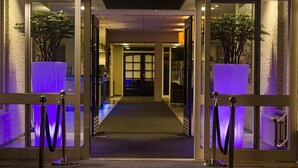 Ein Hotel Eingang mit Glastür, Teppichweg und je einer Seite Kübelpflanzen.