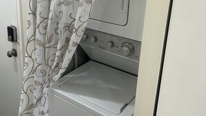 Pequeña lavandería blanca con lavadora y secadora de carga frontal, una puerta con cortina y un panel de control visible.