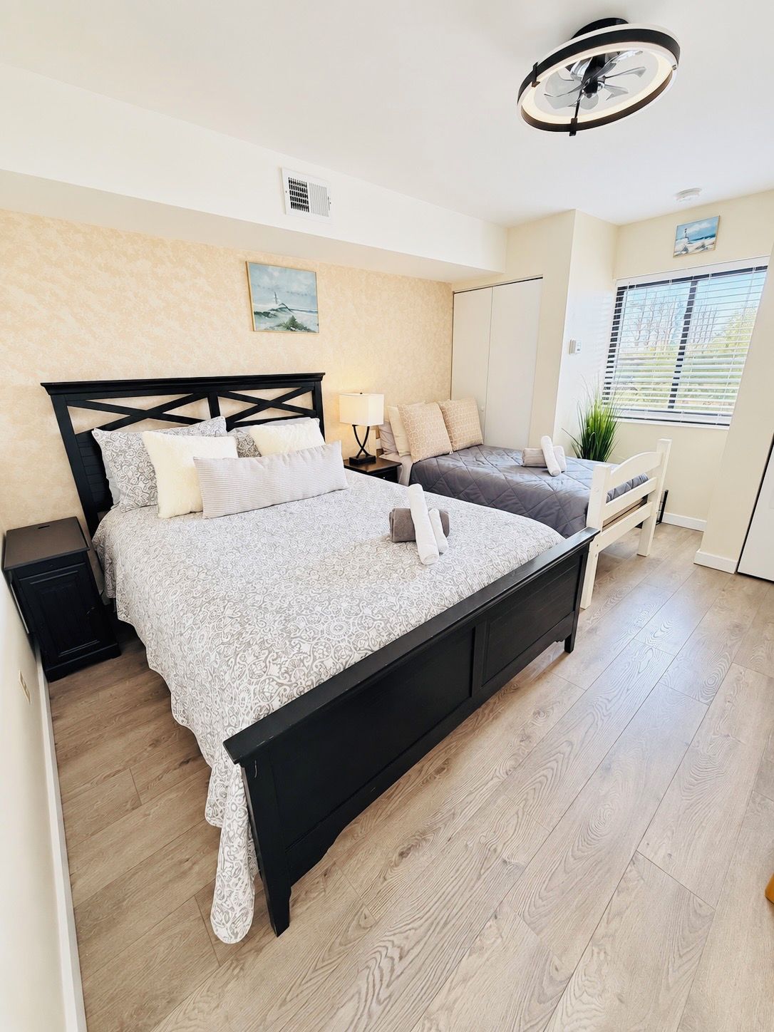 A bedroom with a large bed, a bedside table, a chair, a window with blinds, and a ceiling fan.