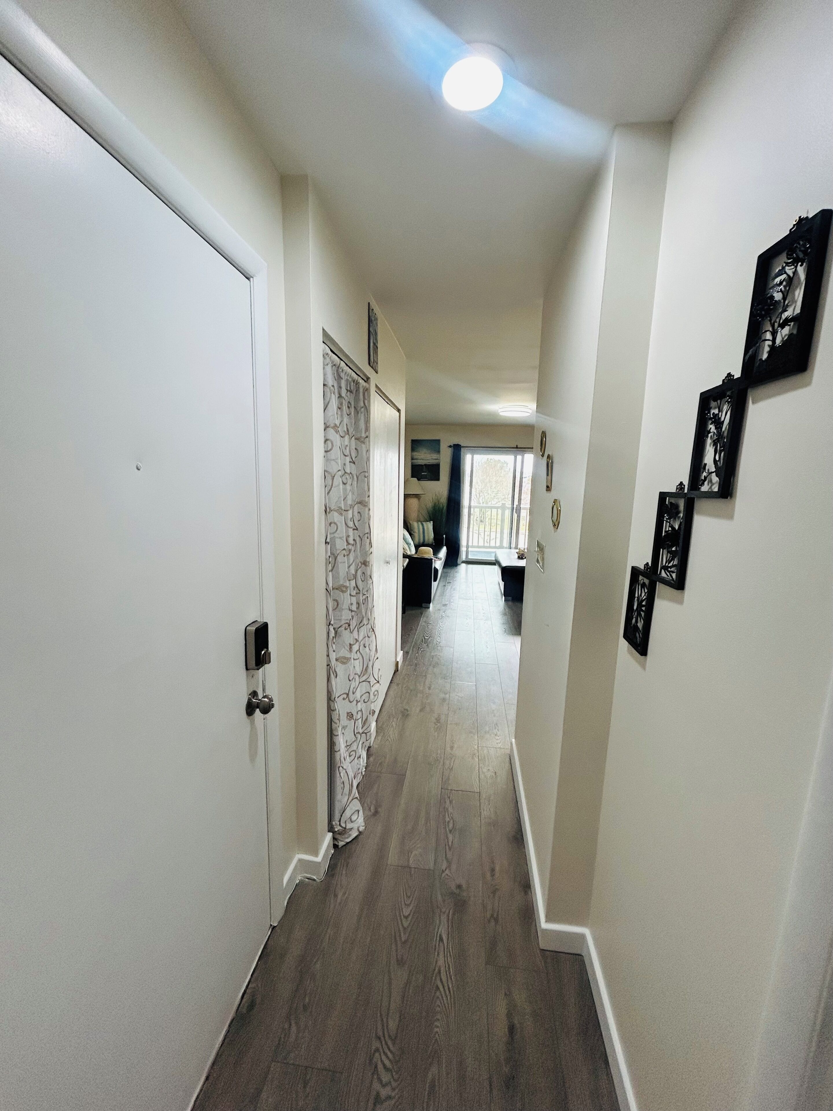Un couloir avec un plancher en bois, des murs blancs et une porte sur le côté gauche. Des tableaux encadrés ornent le mur de droite.