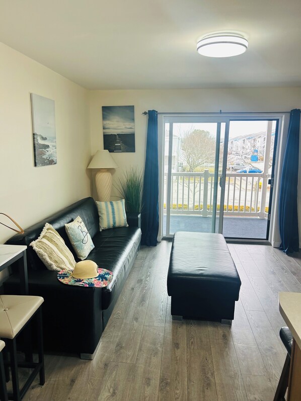 Uma sala moderna com sofá de couro preto, piso de madeira, mesa de jantar e vista para uma varanda com guarda-corrimão.