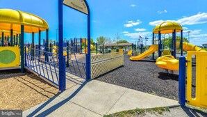Un parque infantil con estructuras de juego amarillas y azules, una entrada con puerta azul, y un cielo despejado.