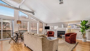 Ein geräumiges Wohnzimmer mit einer großen Couch, zwei Sesseln, einem Couchtisch und einem Kamin. Ein Fernseher ist an der Wand montiert und ein großes Fenster sorgt für natürliches Licht.
