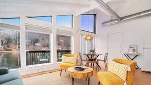 Ein Zimmer mit großen Fenstern und Blick auf ein Gewässer, einem Holztisch, zwei gelben Sesseln und einer Hängeleuchte aus Weidengeflecht.
