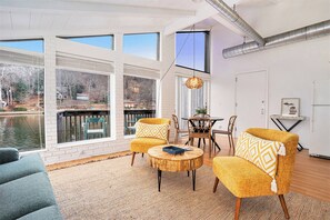 Ein Zimmer mit großen Fenstern und Blick auf ein Gewässer, einem Holztisch, zwei gelben Sesseln und einer Hängeleuchte aus Weidengeflecht.