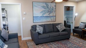 Un salon avec un canapé gris, deux fauteuils, un tapis à motifs et un tableau représentant une scène de plage.