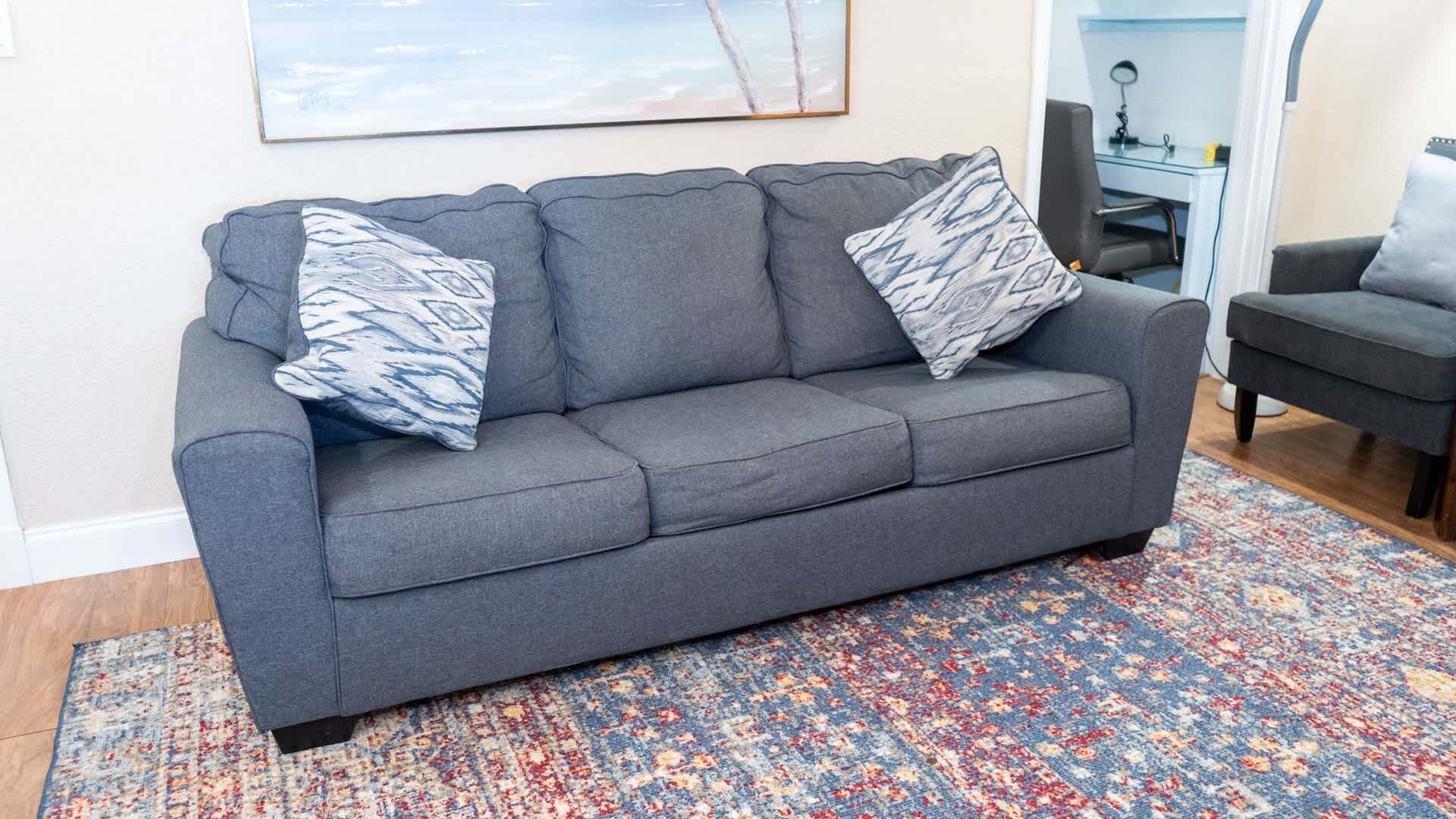 Een grijze bank met gekleurde kussens, een vloerkleed met patroon, en een bureau met stoel op de achtergrond.