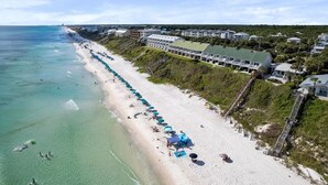 Vue aérienne d’un complexe hôtelier en bord de mer, avec une longue rangée de parasols, une plage de sable et une série de bâtiments dotés d’un toit végétalisé.