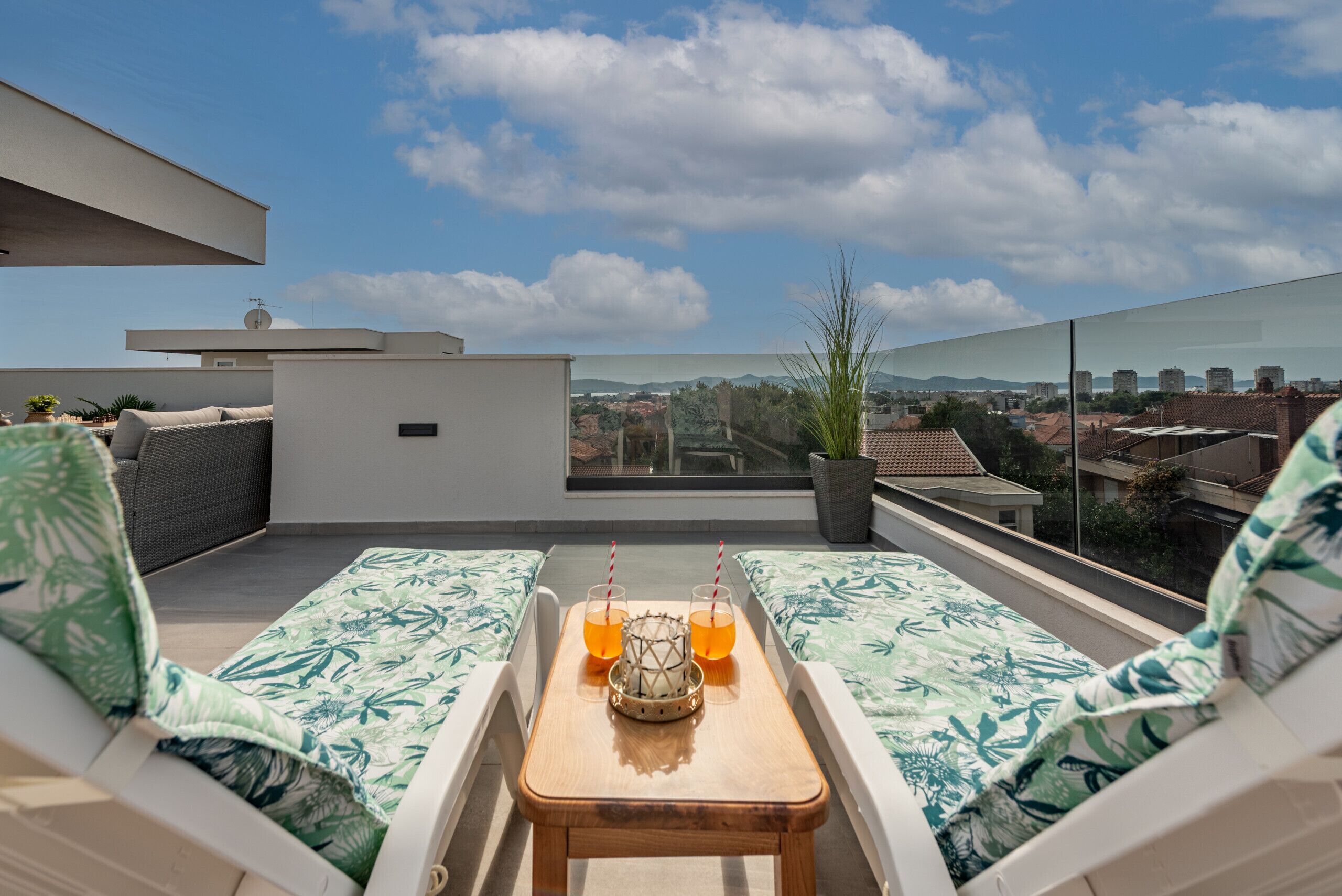 Una terraza en la azotea con dos zonas de estar acolchadas, una mesa de madera con bebidas y vistas a la ciudad.