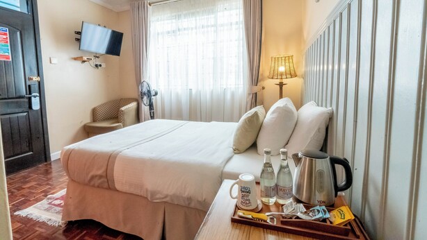 Habitació d'hotel amb un llit, un televisor penjat a la paret, una finestra amb cortines, una tauleta de nit amb un plat de productes de tocador i un radiador.