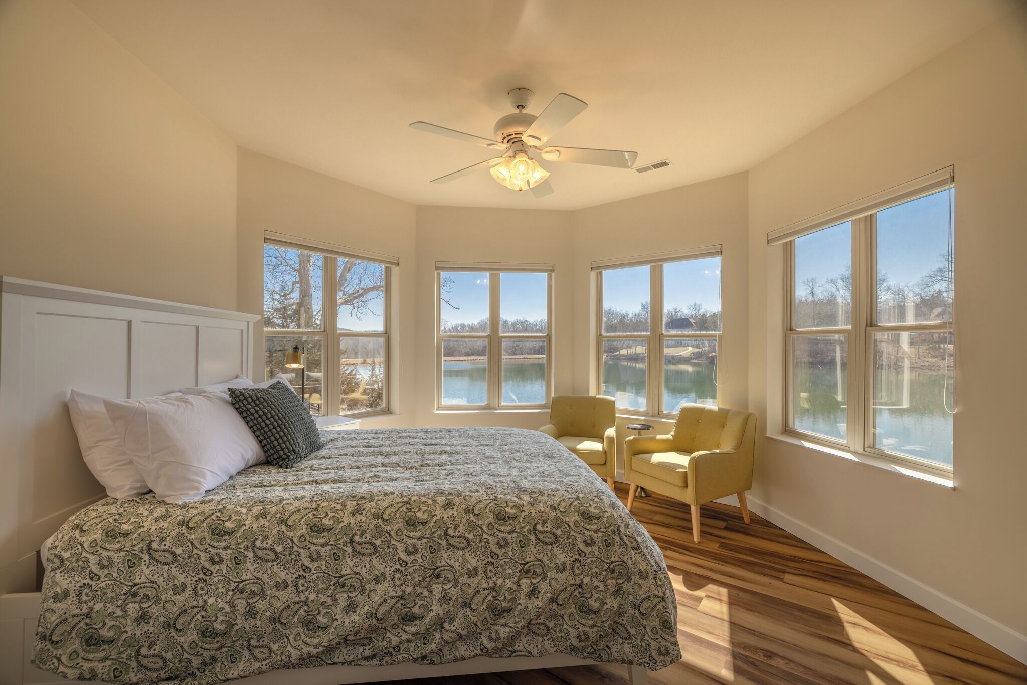 Ein Schlafzimmer mit einem Bett, zwei Stühlen, einem Deckenventilator und großen Fenstern mit Blick auf ein Gewässer.