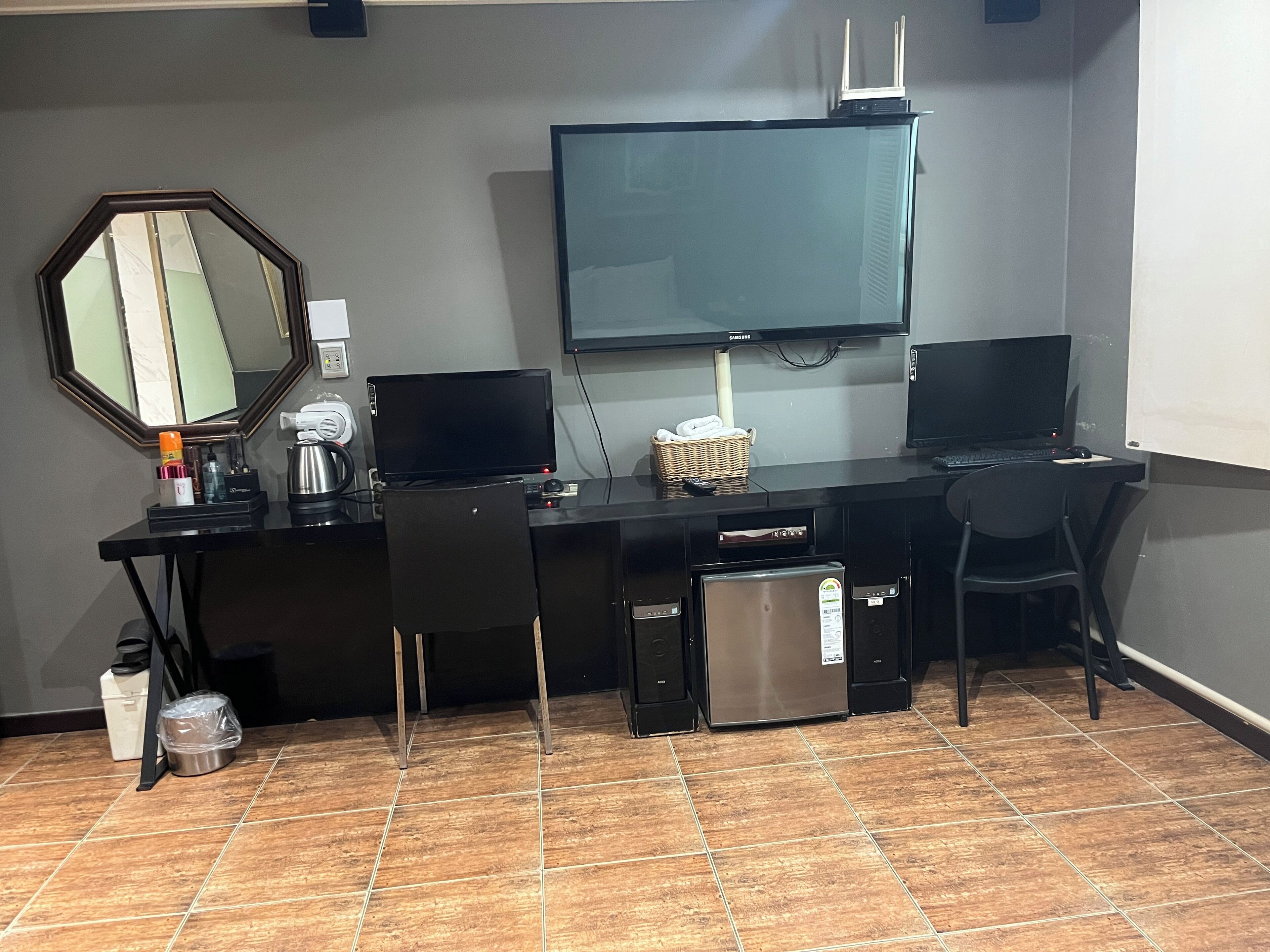 A hotel room with a black desk, two monitors, a refrigerator, a microwave, a coffee maker, a basket, a chair, and a large mirror.