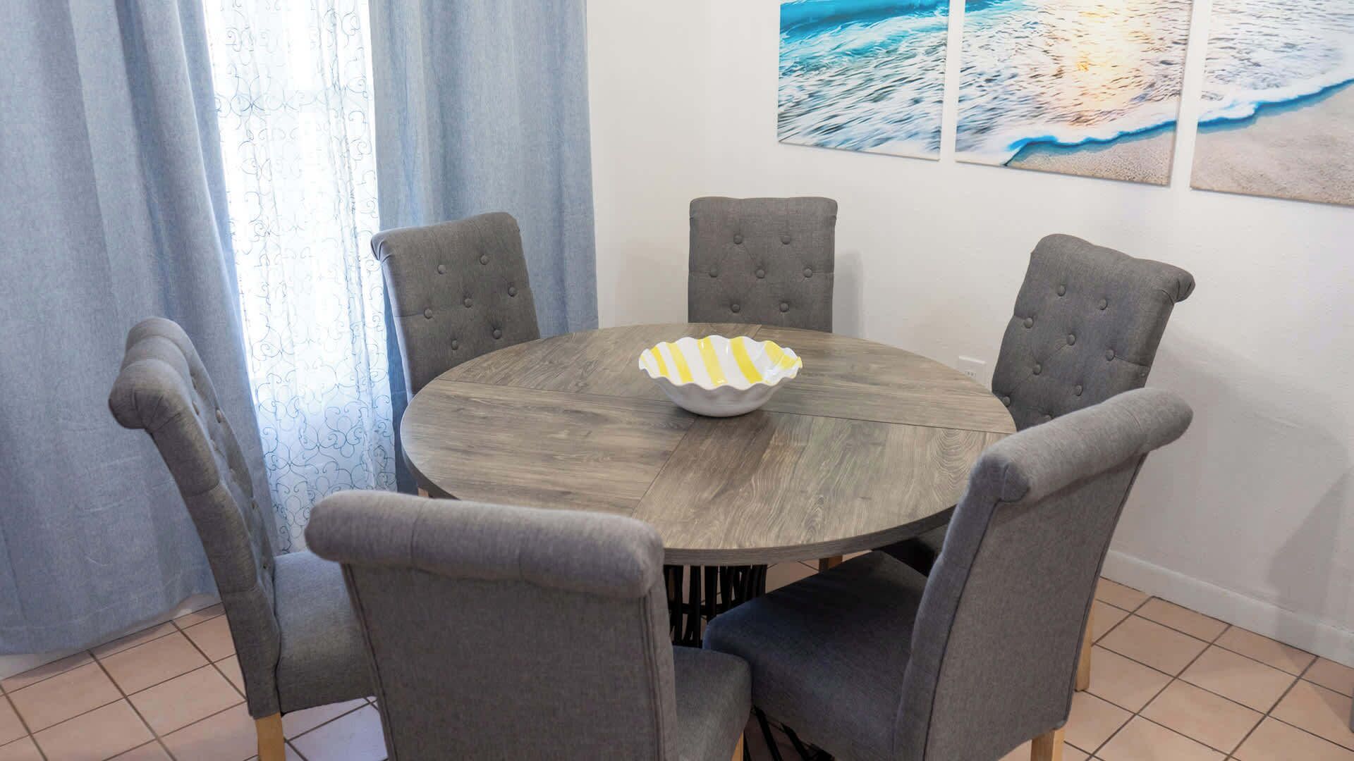 Une table de salle à manger ronde, avec six chaises grises à revêtement rembourré, un plateau en bois et un bol décoratif blanc et jaune.