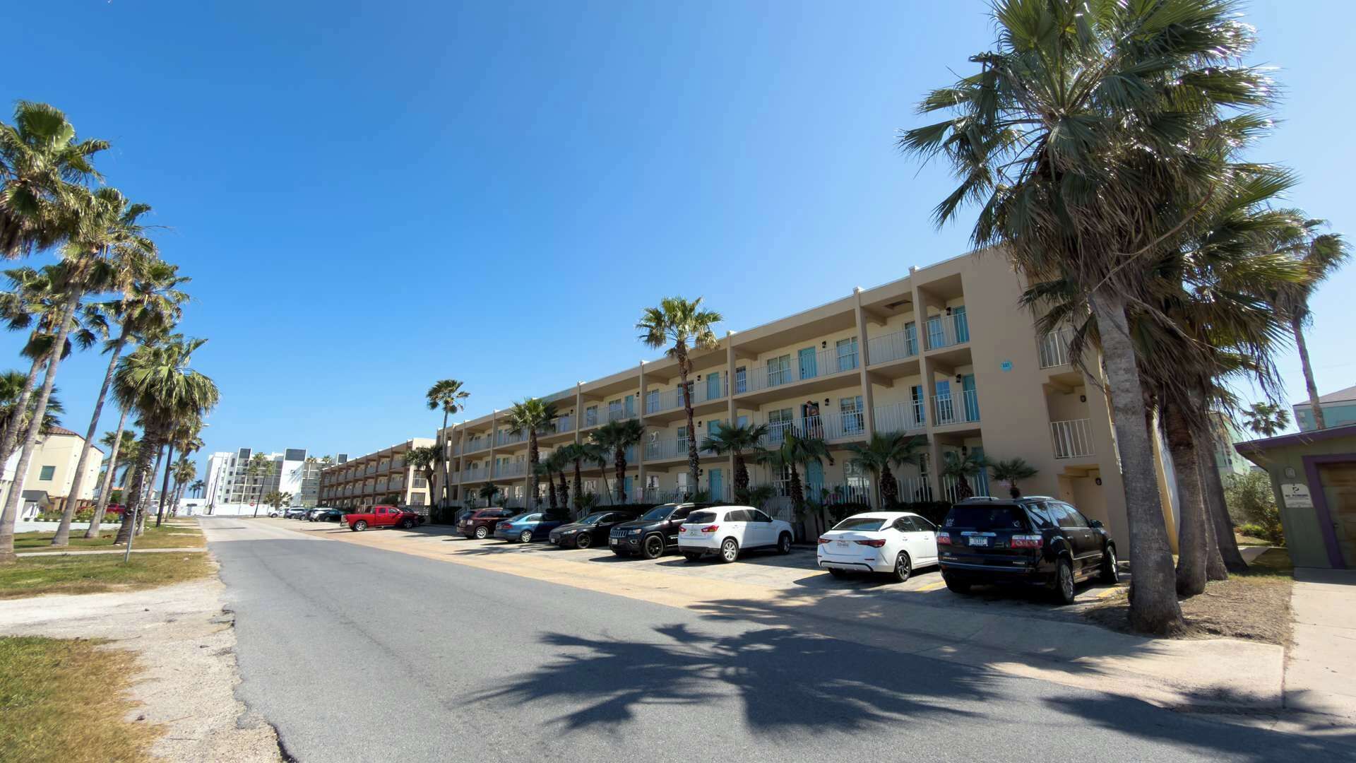 Un hôtel à plusieurs étages, avec une rangée de voitures garées et des palmiers bordant la rue.